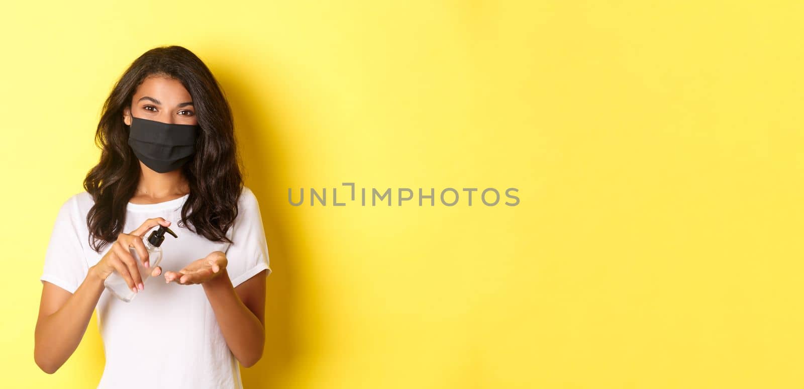 Concept of covid-19, social distancing and lifestyle. Image of african-american girl in face mask using hand sanitizer, standing over yellow background by Benzoix