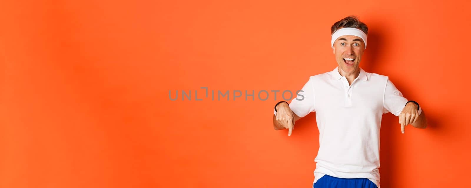 Concept of workout, sports and lifestyle. Portrait of happy middle-aged man in gym uniform, pointing fingers down and smiling amazed, showing promo offer, standing over orange background.