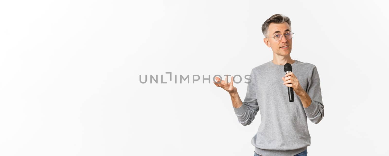 Portrait of handsome middle-aged man in glasses and gray sweater, making speech, talking with microphone and explaining something, standing over white background.