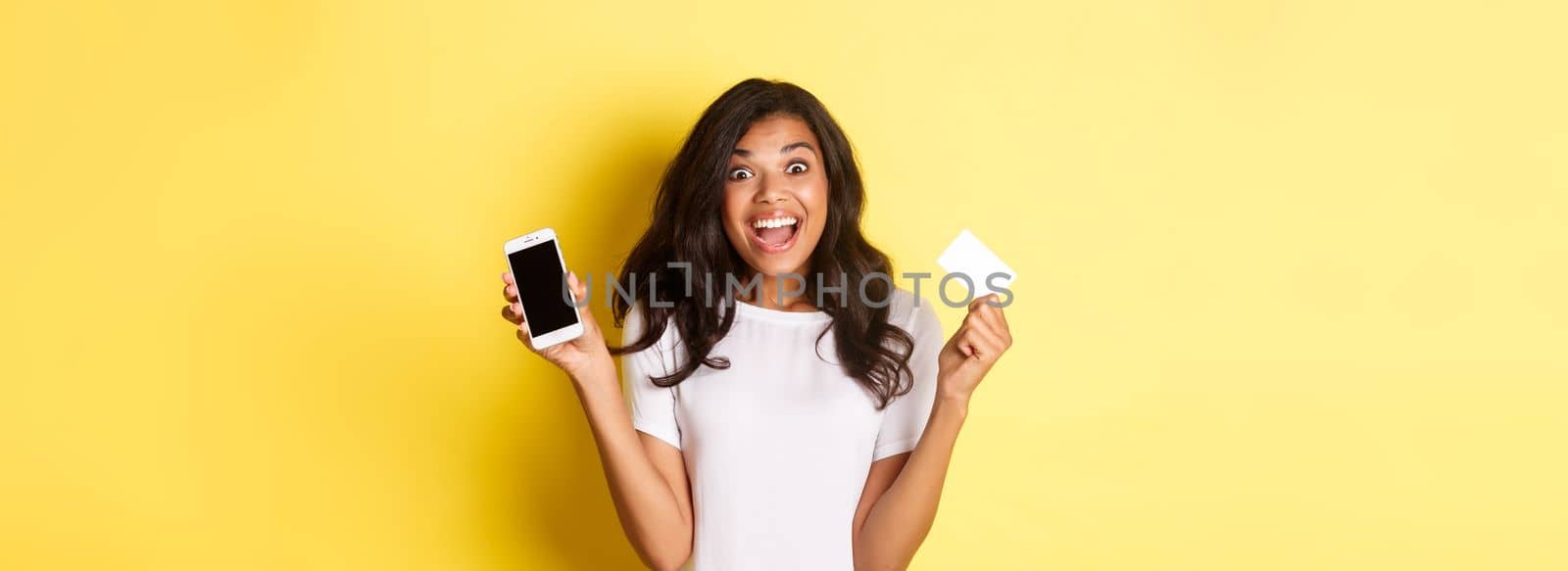Image of excited african-american female model, showing smartphone screen and credit card, standing over yellow background by Benzoix