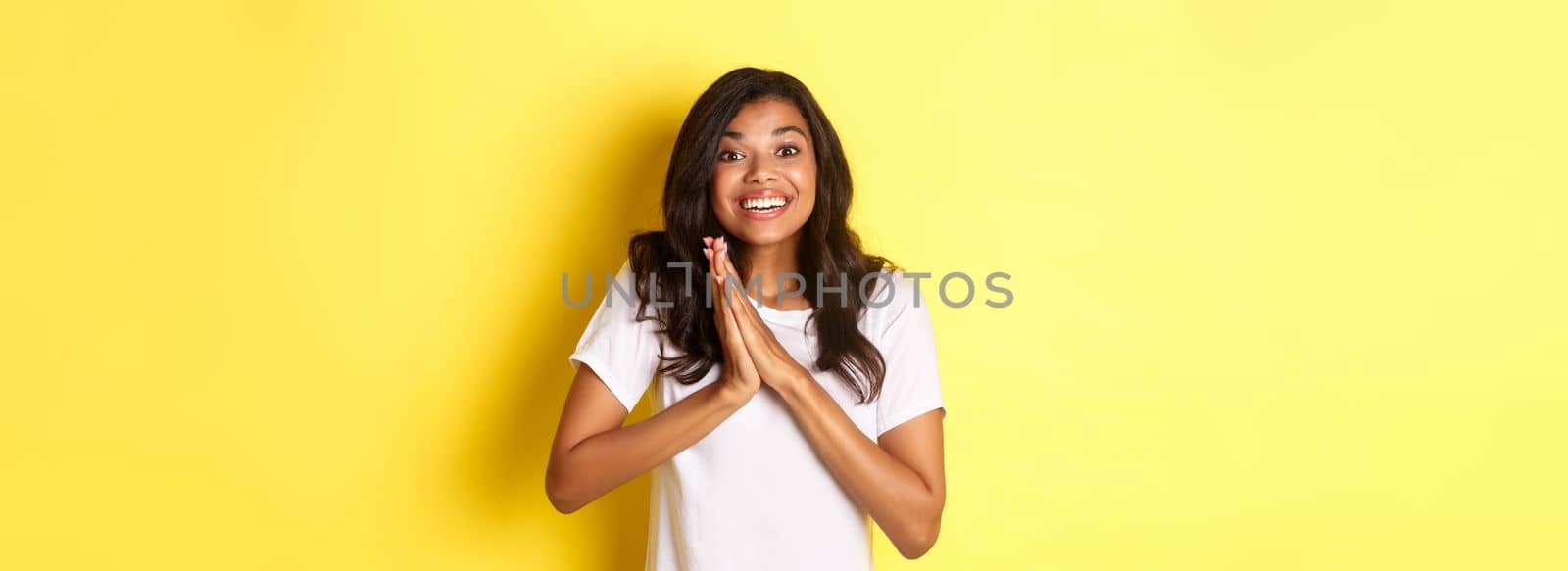 Image of happy and pleased african-american girl, clap hands and looking excited, thanking for something, standing over yellow background by Benzoix