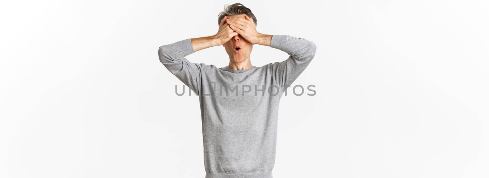 Image of excited middle-aged guy in grey sweater, cover eyes with hands and looking amazed while waiting for surprise, playing hide-n-seek, standing over white background.
