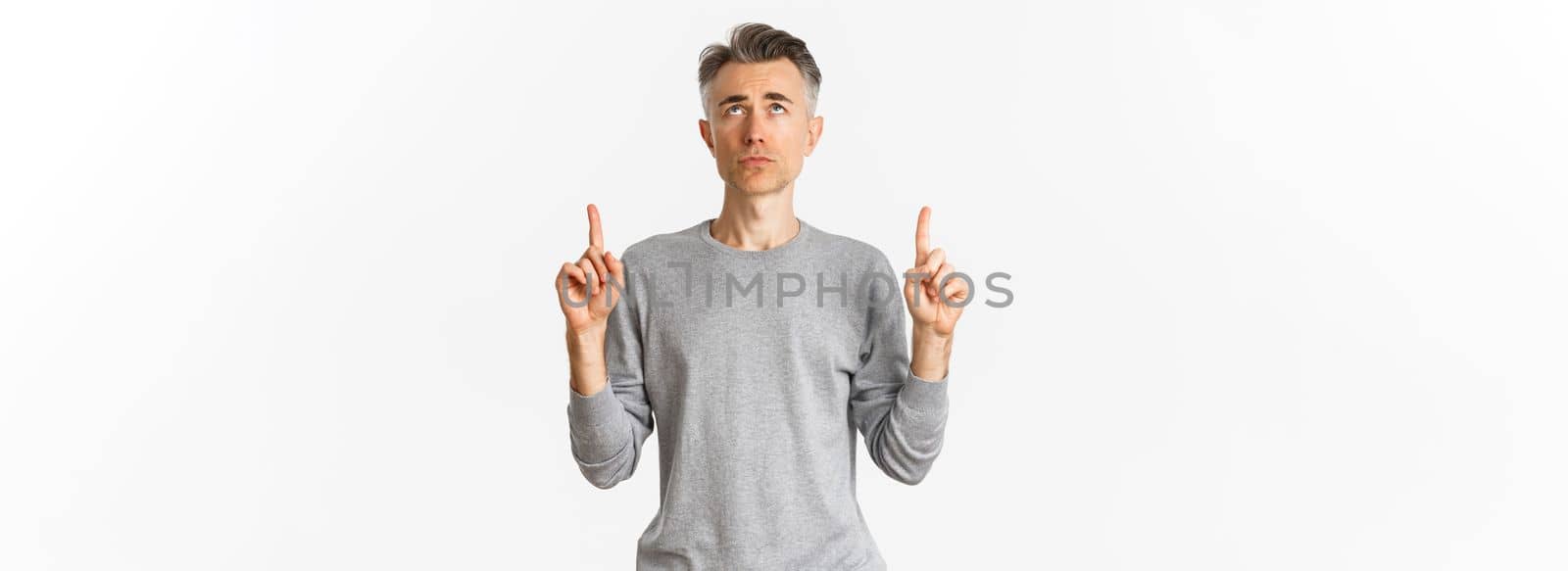 Portrait of gloomy and hesitant middle-aged man, looking troubled and pointing fingers up, standing upset over white background.
