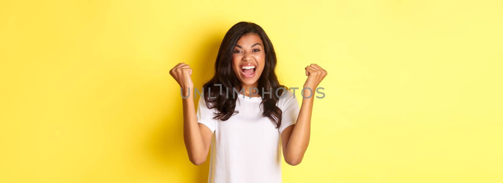 Image of successful african-american girl, feeling lucky, making fist pump sign and saying yes, triumphing and shouting for joy, standing over yellow background by Benzoix