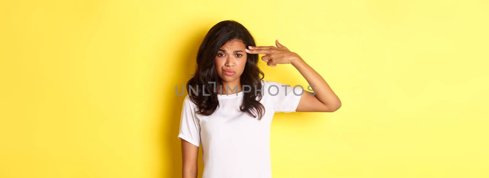 Portrait of skeptical and bothered african-american woman, making finger gun sign over head and smirking unamused, standing over yellow background.