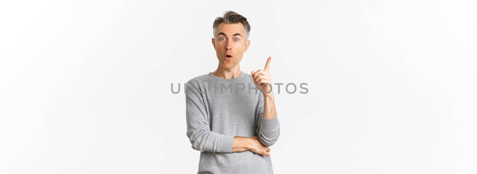 Portrait of thoughtful adult male model with short grey hair, raising finger up, having an idea, say suggestion, standing over white background.