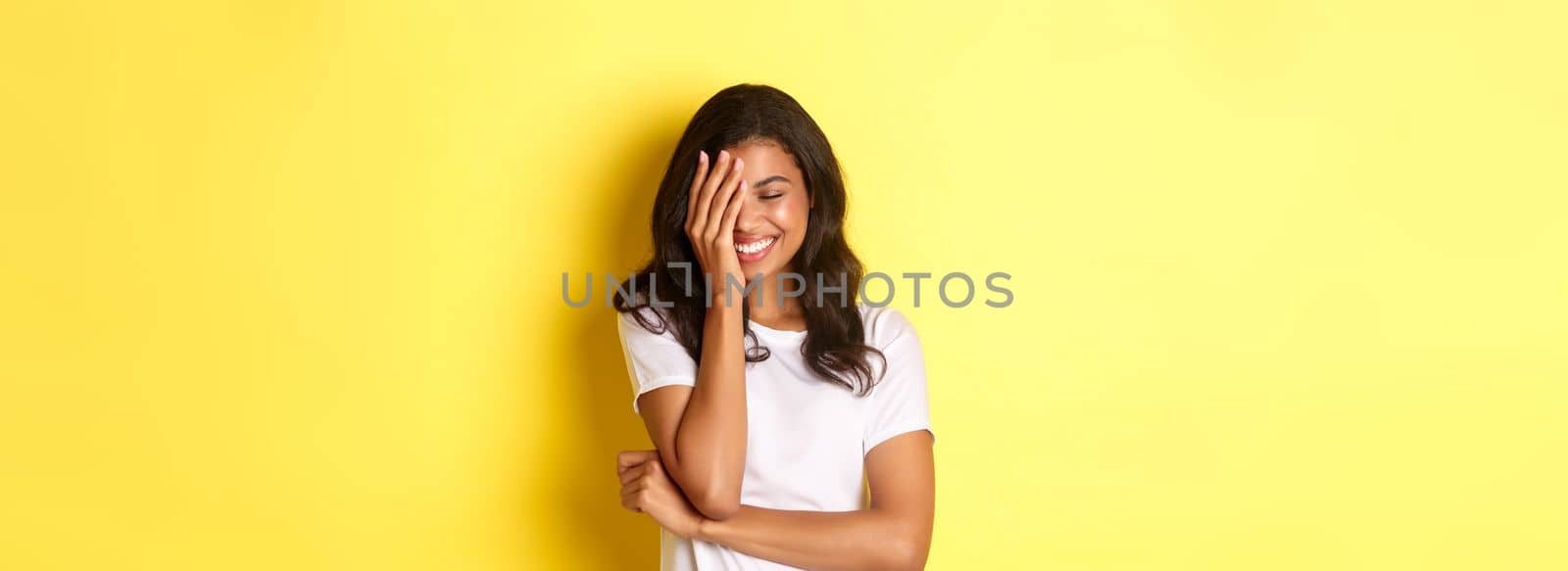 Portrait of beautiful and happy african-american girl, silly laughing and covering her face with hand, standing over yellow background.