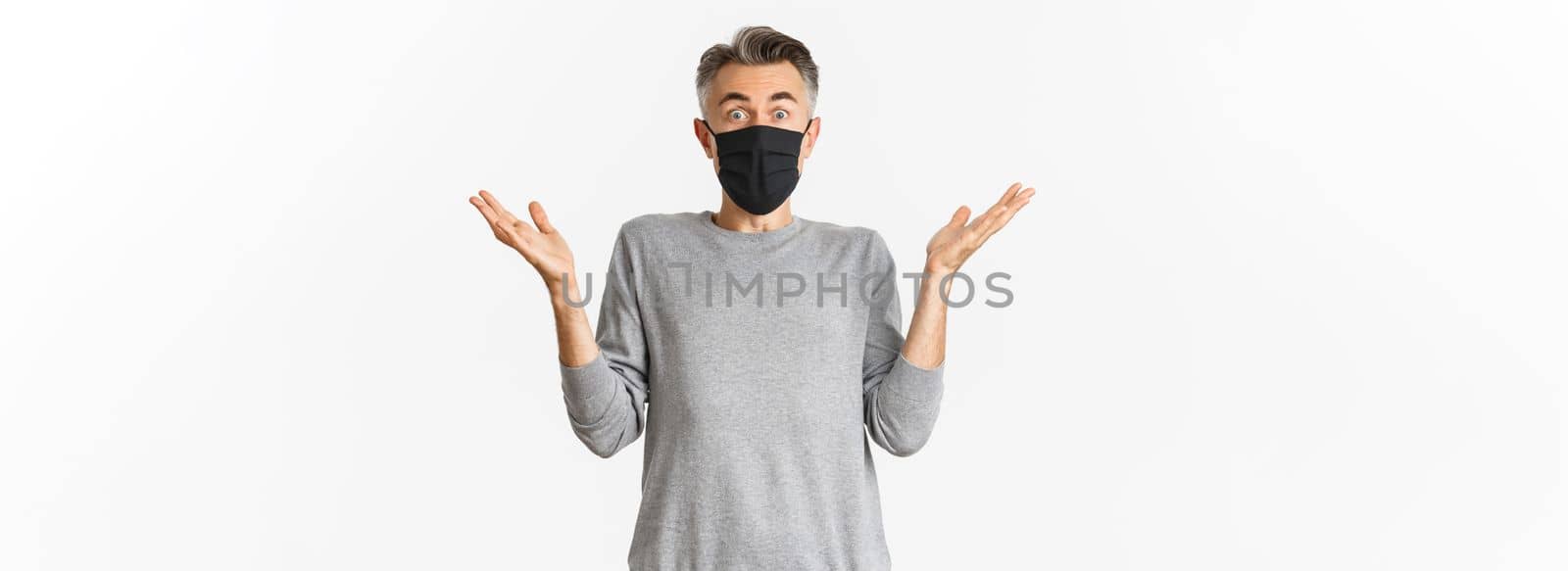 Covid-19, pandemic and social distancing concept. Portrait of handsome middle-aged man in black medical mask looking surprised, reacting amazed, standing over white background.