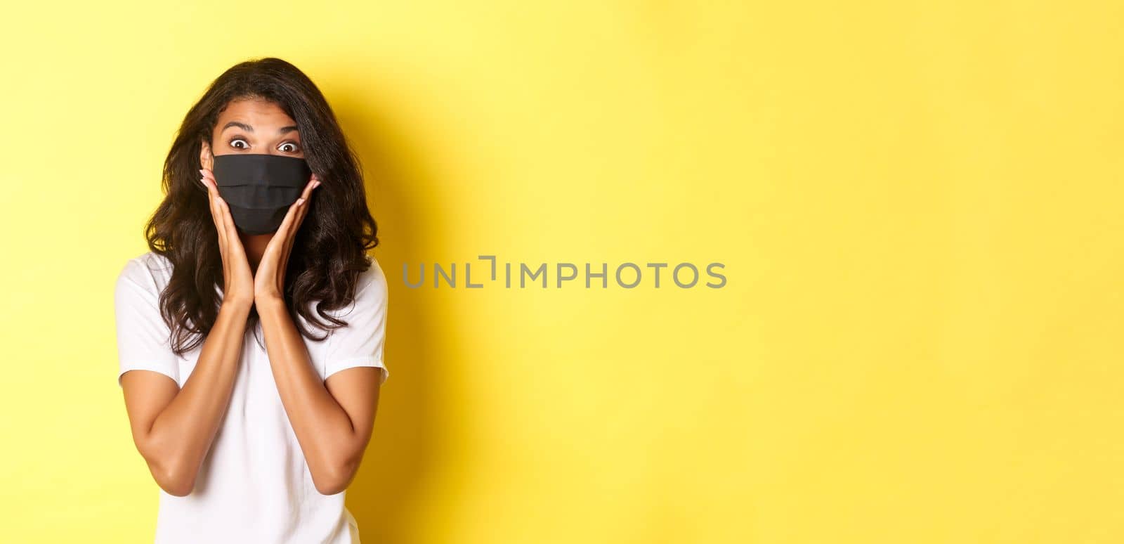 Concept of coronavirus, pandemic and lifestyle. Portrait of excited african-american woman in face mask, looking amazed at something cool, standing over yellow background by Benzoix