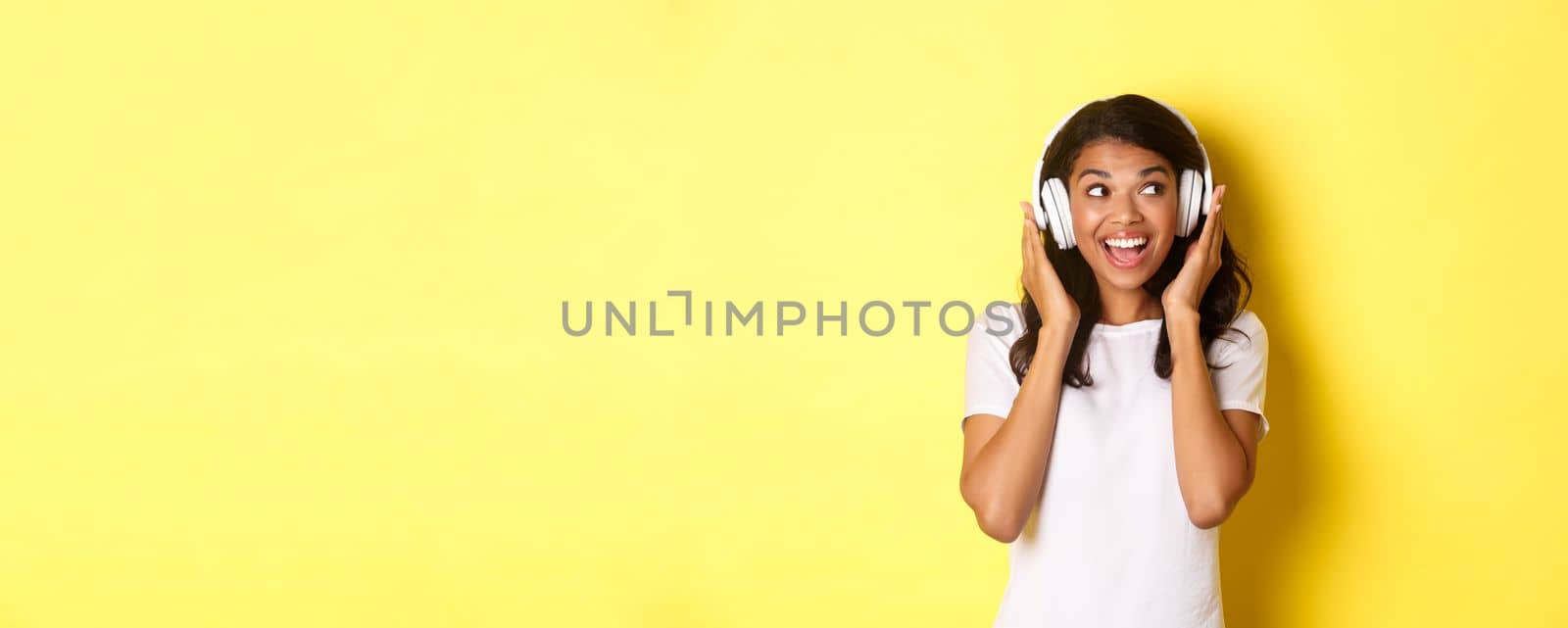 Portrait of cute african-american girl, smiling while listening to music in headphones, looking aside happy, standing over yellow background by Benzoix