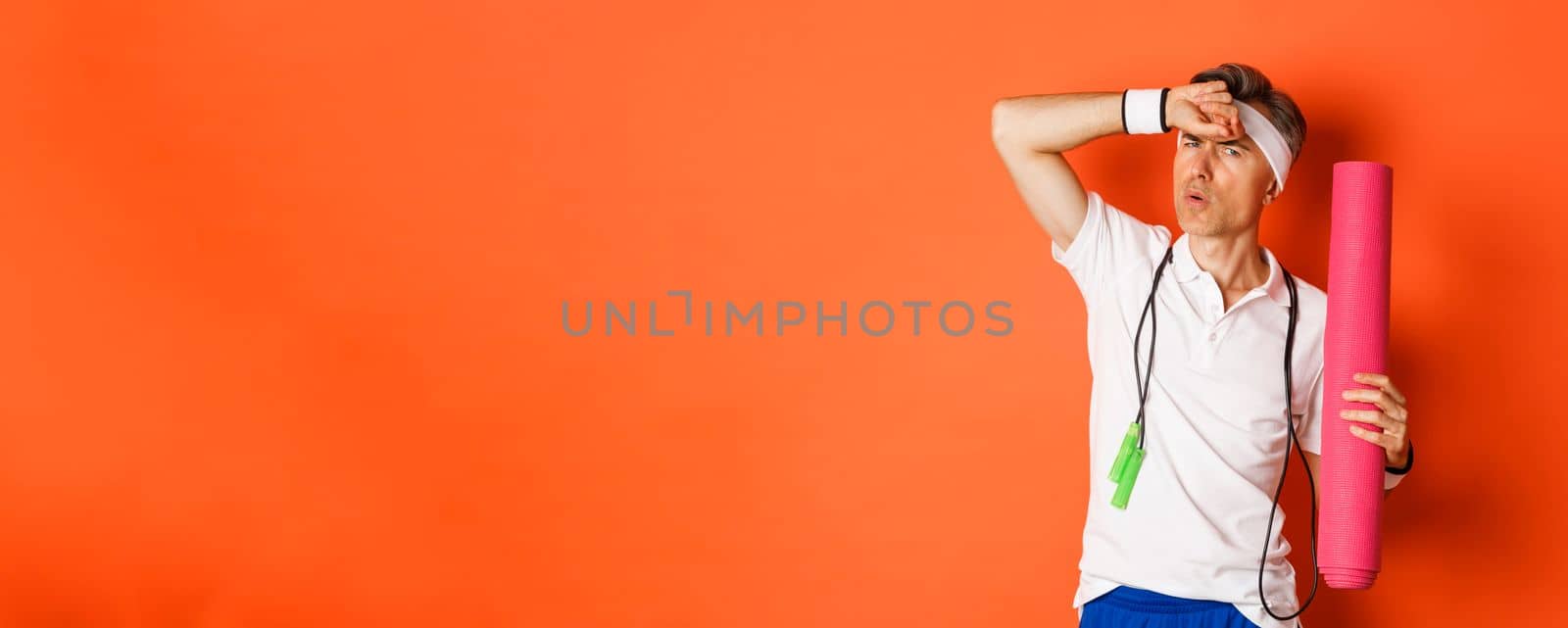 Concept of workout, gym and lifestyle. Image of handsome middle-aged man, tired after fitness exercises, holding skipping rope and yoga mat, wiping sweat off forehead, orange background by Benzoix