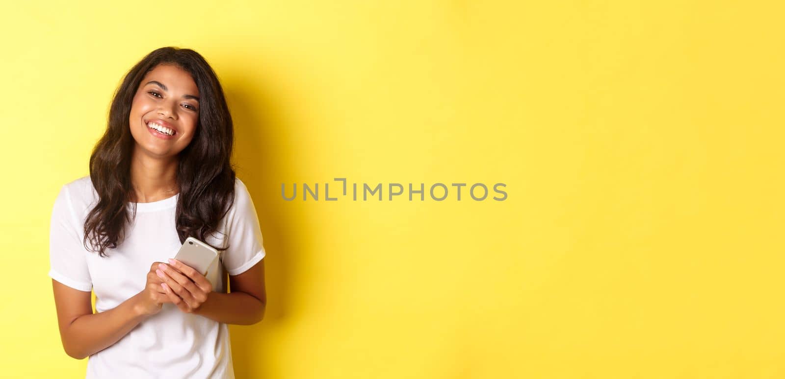 Portrait of beautiful african-american woman, smiling happy and holding smartphone, standing over yellow background by Benzoix