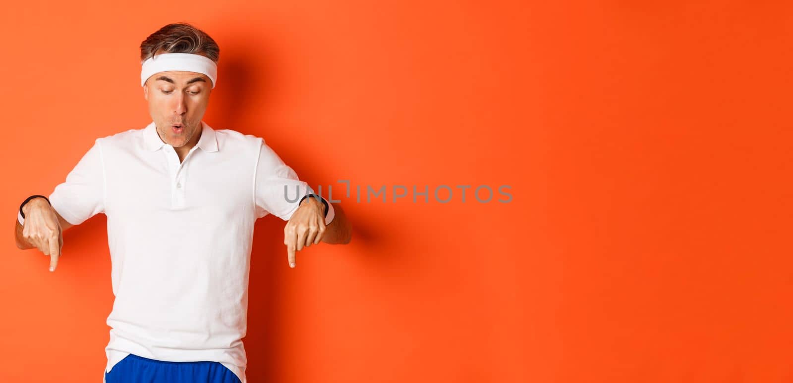 Concept of workout, sports and lifestyle. Amazed and excited middle-aged sportsman in fitness clothing, pointing fingers down and looking amused at logo, standing over orange background.