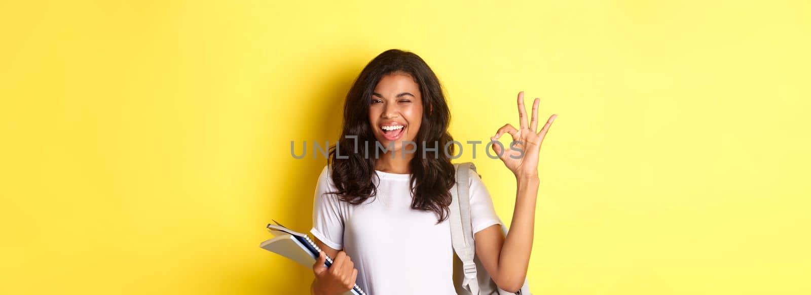 Portrait of confident african-american female student, showing okay sign and winking, approve something good, standing with notebooks and backpack for college, yellow background.