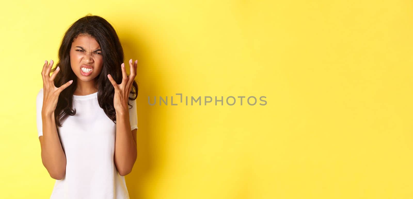 Image of frustrated and angry african-american girl, grimacing and shaking hands mad, standing over yellow background.