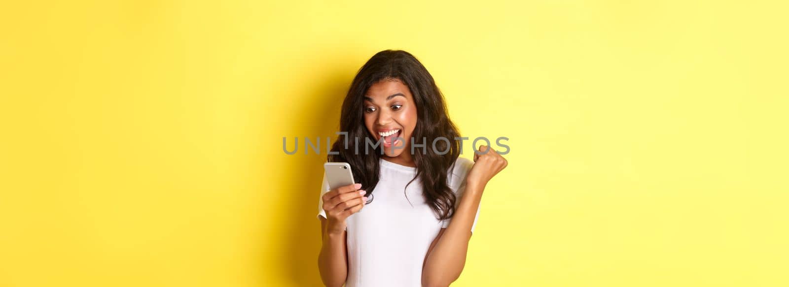 Portrait of beautiful african-american girl, rejoicing and looking at mobile phone, found something amazing online, standing over yellow background.