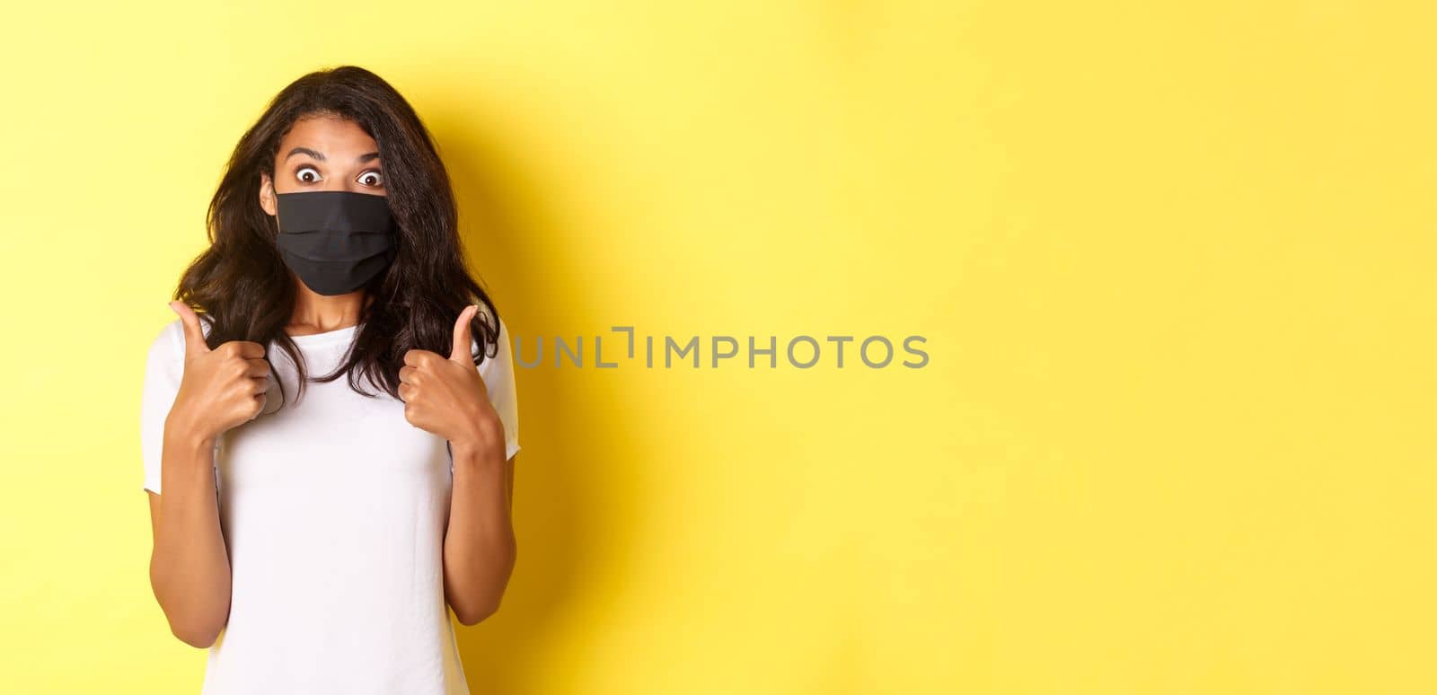 Concept of coronavirus, pandemic and lifestyle. Portrait of beautiful african-american girl in black face mask, showing thumbs-up and looking amazed, recommending something, yellow background.