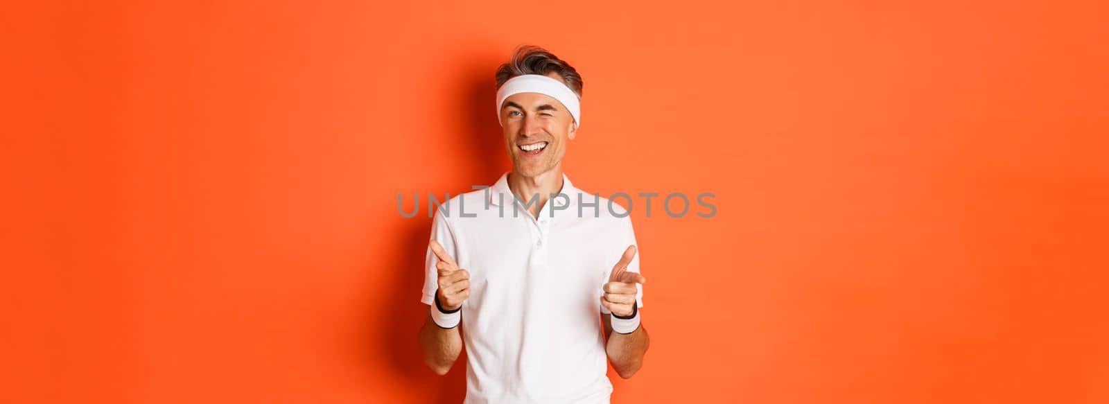 Concept of sport, fitness and lifestyle. Portrait of confident, handsome middle-aged man in workout uniform, pointing fingers at camera and smiling, inviting to gym, standing over orange background by Benzoix