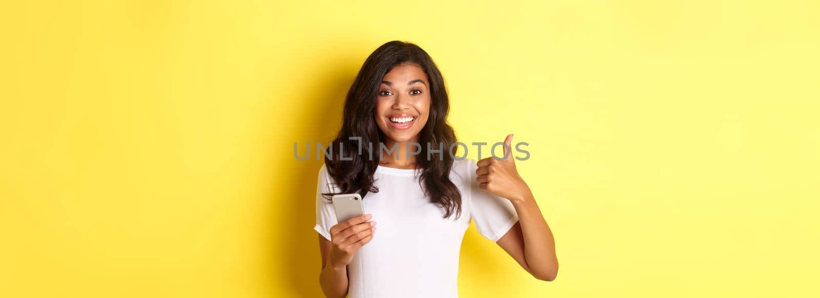 Portrait of young good-looking african american woman, showing thumbs-up while using mobile phone app, smiling pleased, standing over yellow background.