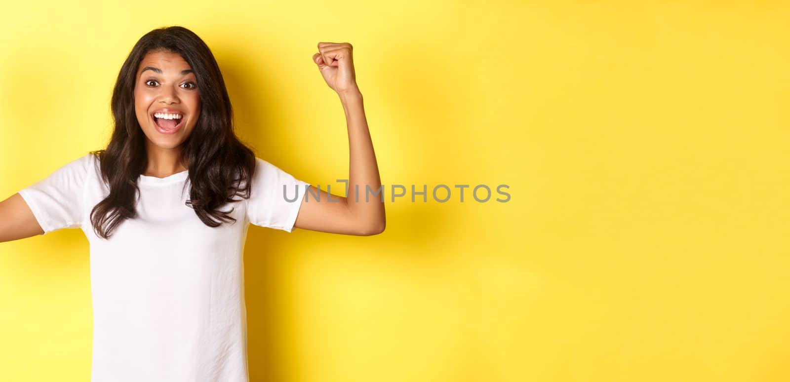 Image of happy african-american girl achieve goal and celebrating victory, raising hands up and smiling pleased, looking satisfied, standing over yellow background.