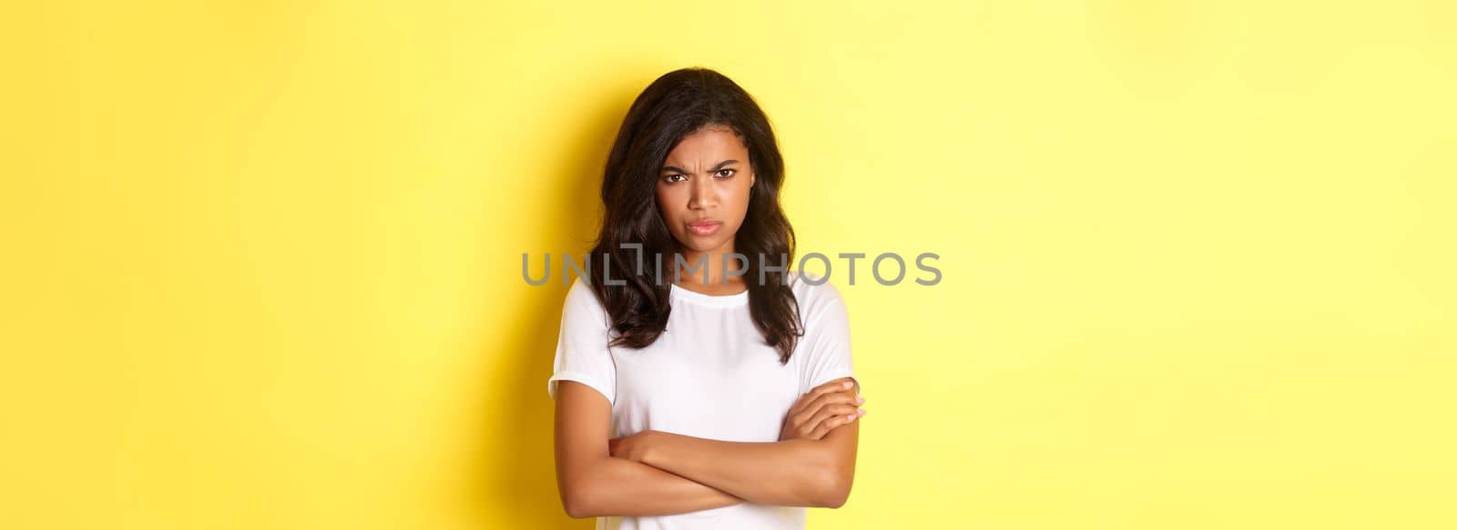 Image of cute african-american girl feeling angry, cross arms on chest and frowning, mad at you, standing over yellow background.