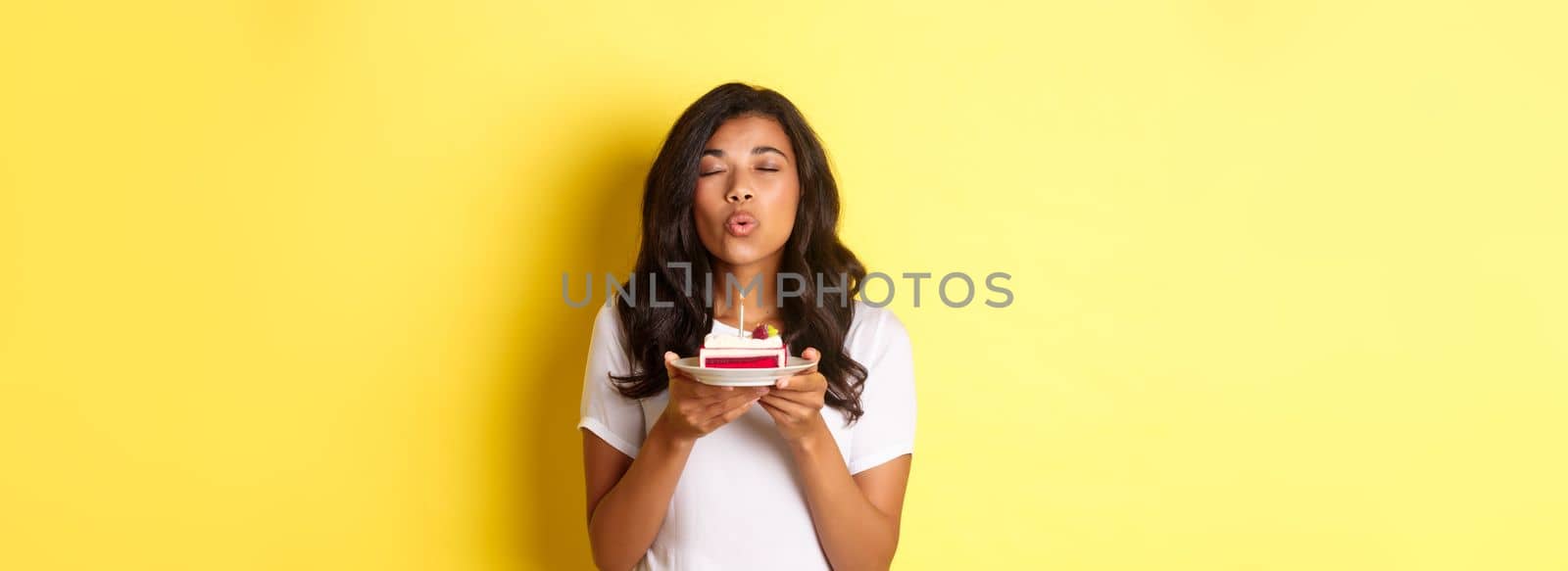 Portrait of beautiful african-american girl celebrating birthday, blowing lit candle on b-day cake and making a wish, standing over yellow background by Benzoix