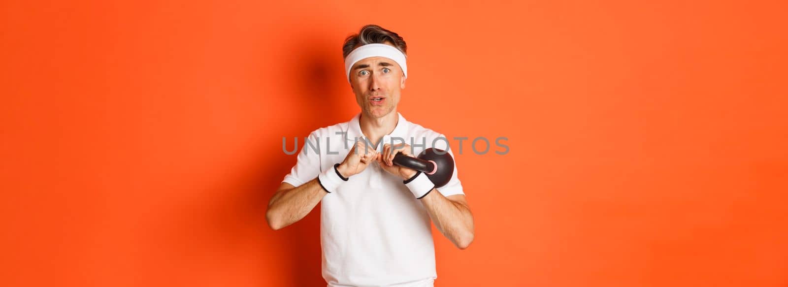 Concept of workout, gym and lifestyle. Close-up of weak and slim middle-aged fitness guy, doing sport exercises with kettlebell, standing over orange background by Benzoix