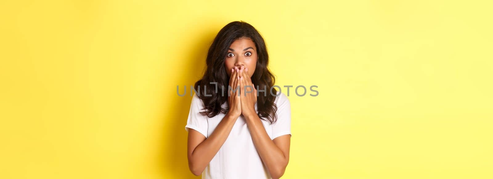 Portrait of startled african-american woman, gasping and covering mouth with hands, looking shocked at camera, standing over yellow background for copy space.