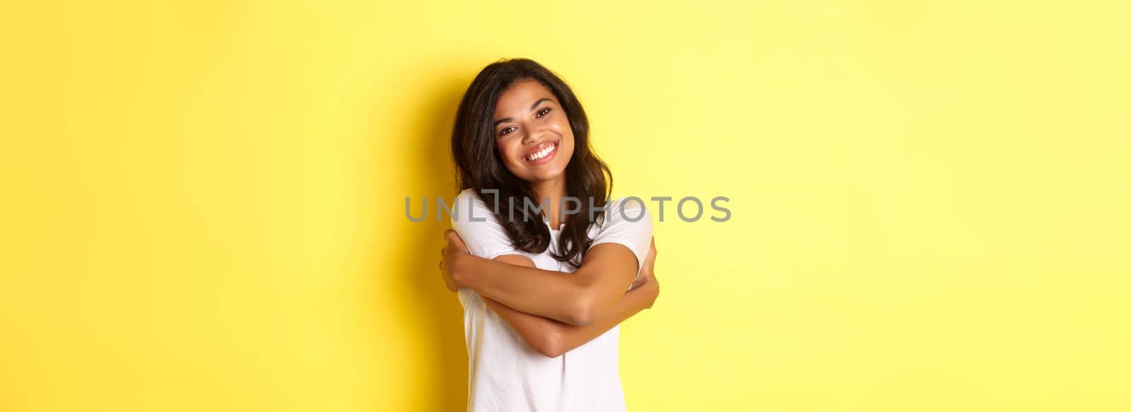 Portrait of cheerful african american woman, hugging herself and smiling pleased, standing over yellow background.