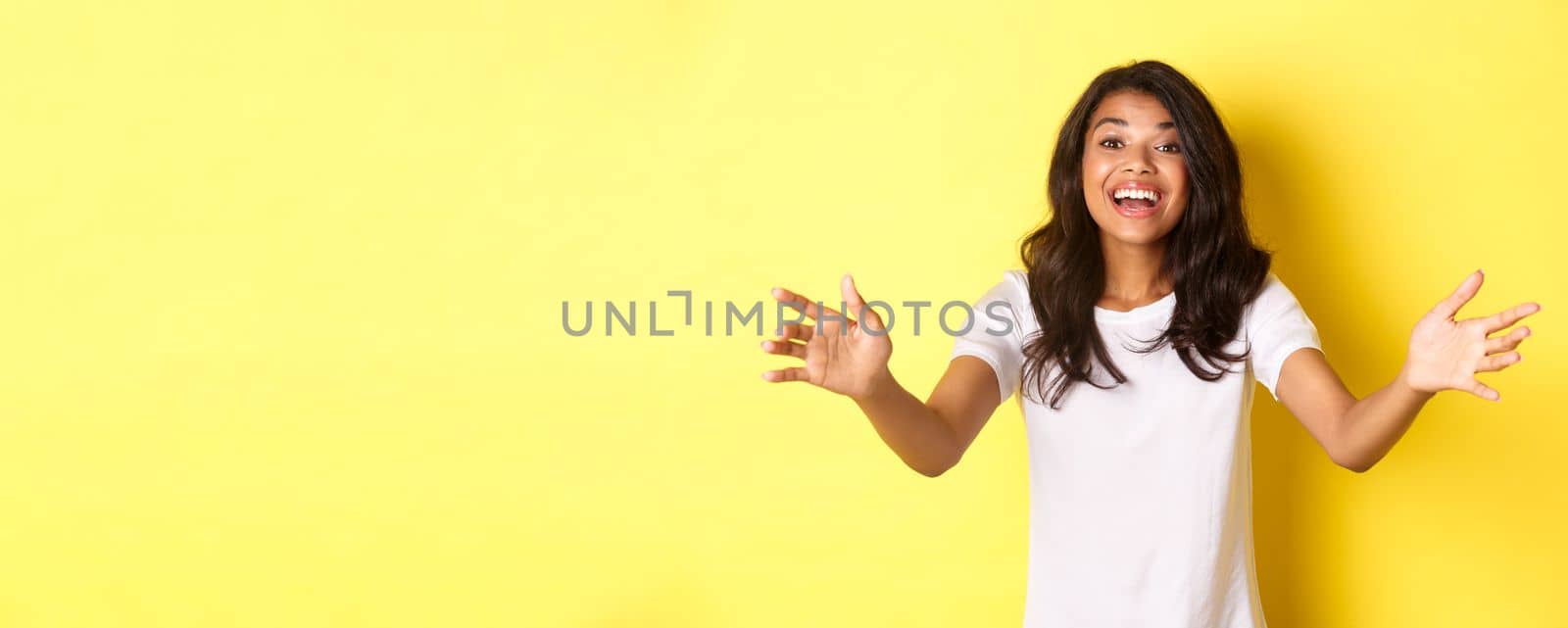 Portrait of beautiful and friendly african-american girl, reaching hands to you, wanting to hold or take something, hugging person with pleased smile, standing over yellow background.