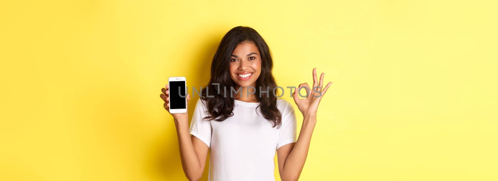 Portrait of good-looking african-american girl in white t-shirt, showing okay sign and smartphone app, standing over yellow background by Benzoix