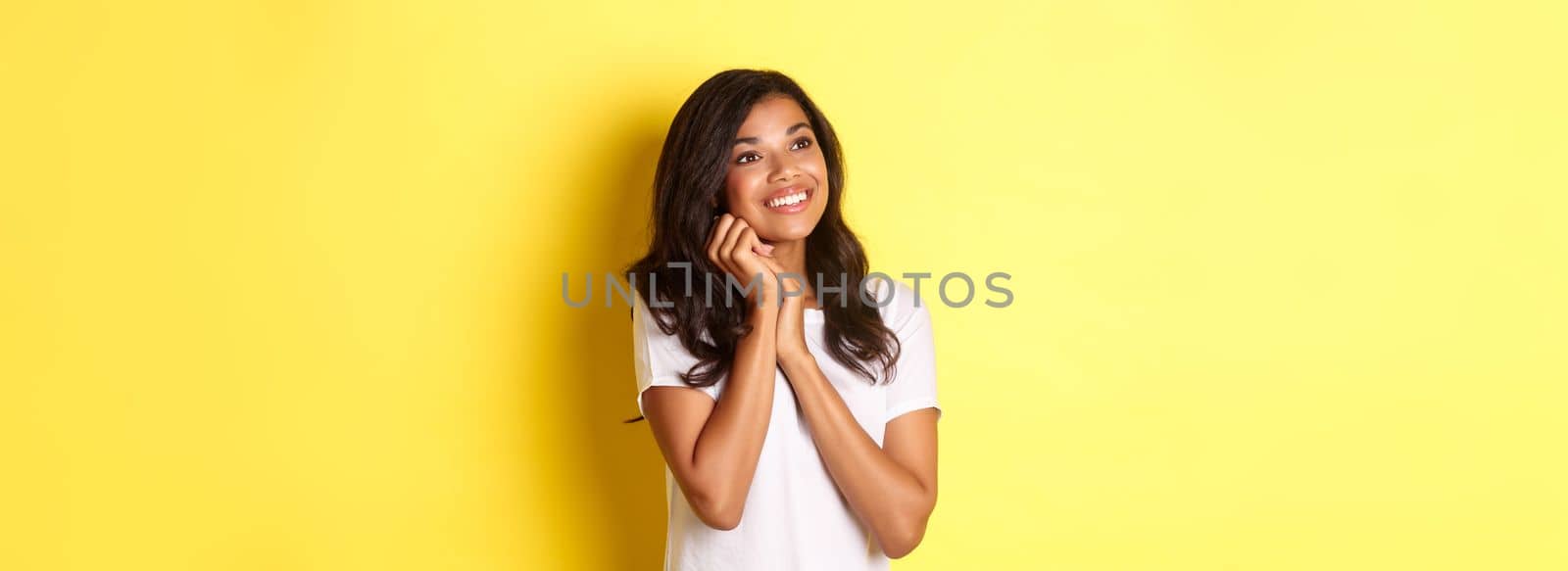 Portrait of dreamy and beautiful african-american girl, looking with admiration at upper left corner and smiling, daydreaming over yellow background.