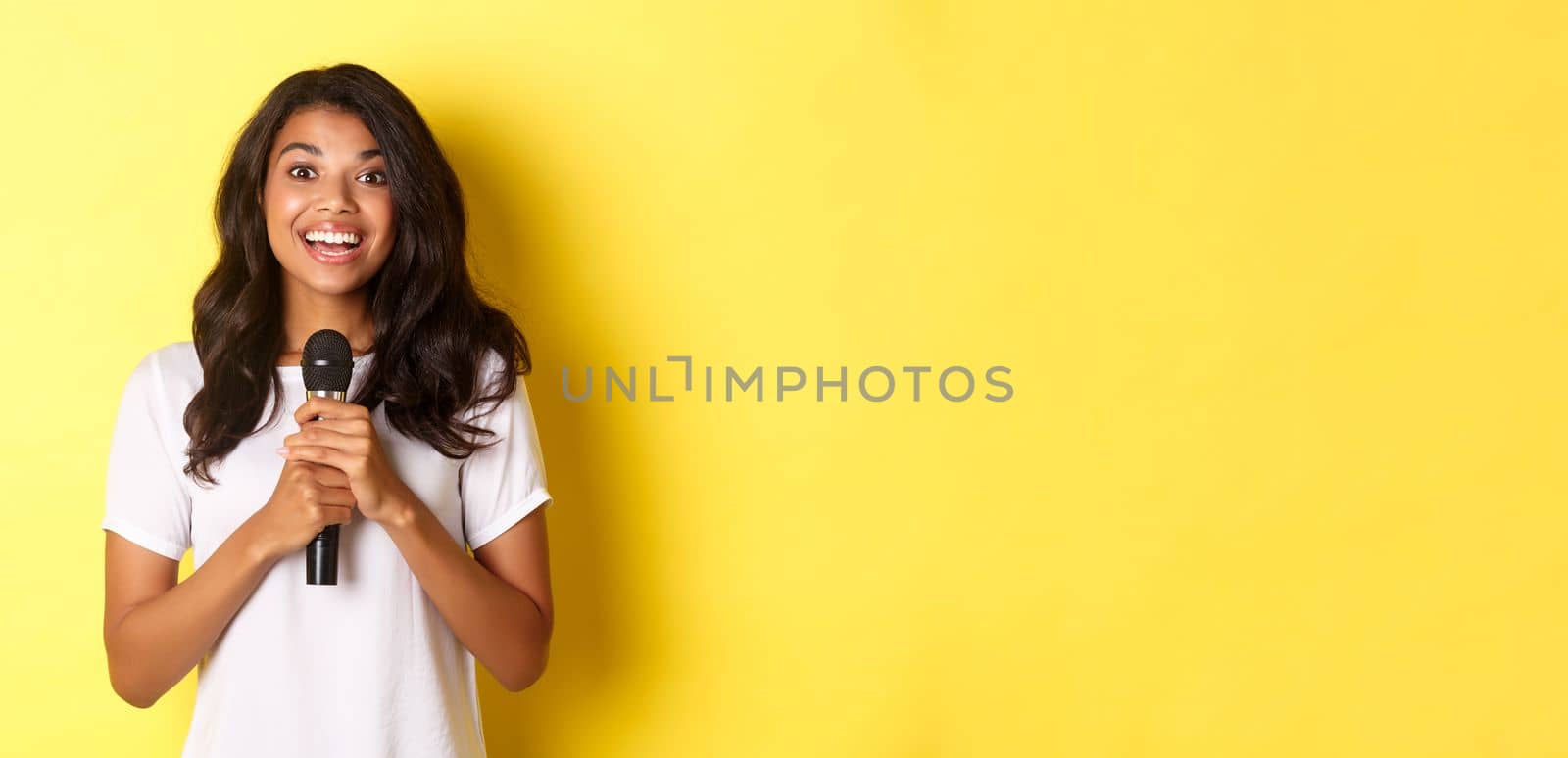 Portrait of happy african-american girl, looking amused while giving speech, holding microphone and smiling at camera, standing over yellow background by Benzoix