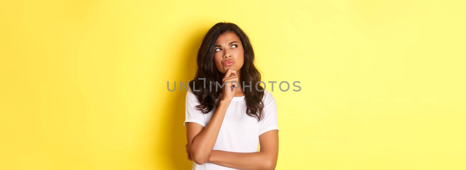 Portrait of attractive african-american woman thinking, making her choice, looking at upper left corner thoughtful and deciding, standing over yellow background.