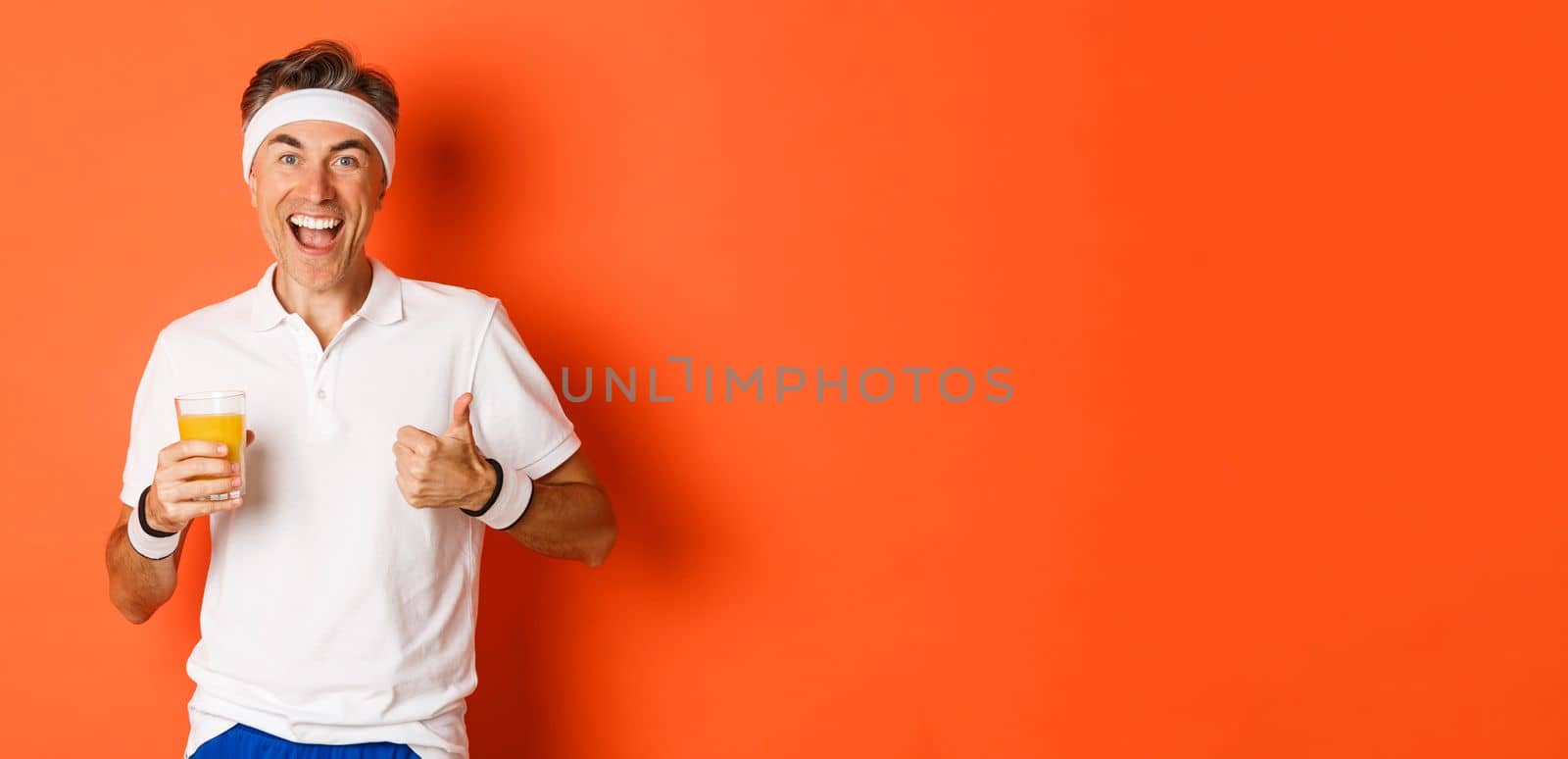 Concept of workout, gym and lifestyle. Image of active and healthy middle-aged sport guy, showing thumbs-up and drinking juice, smiling happy, standing over orange background.