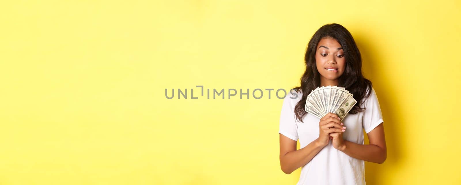 Portrait of attractive african-american woman, looking tempted at money, wanting to buy something, standing over yellow background by Benzoix