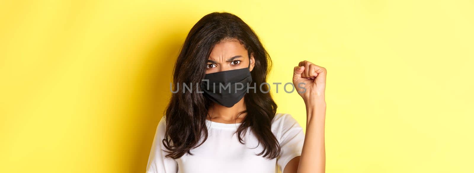 Concept of covid-19, social distancing and lifestyle. Close-up of fierce and confident african-american woman, being a protester in black lives matter movement, showing a fist, wearing face mask.