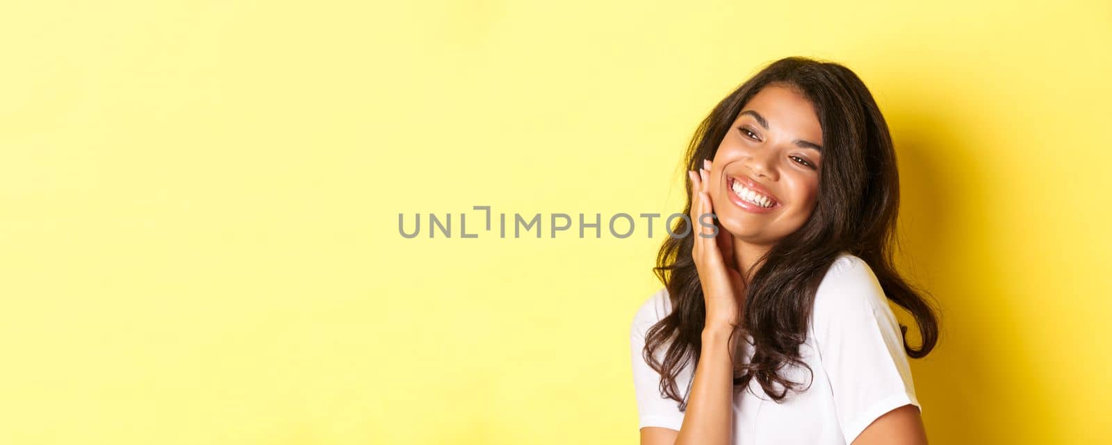 Image of gorgeous african-american woman touching her face, smiling pleased and looking left at copy space, standing over yellow background by Benzoix