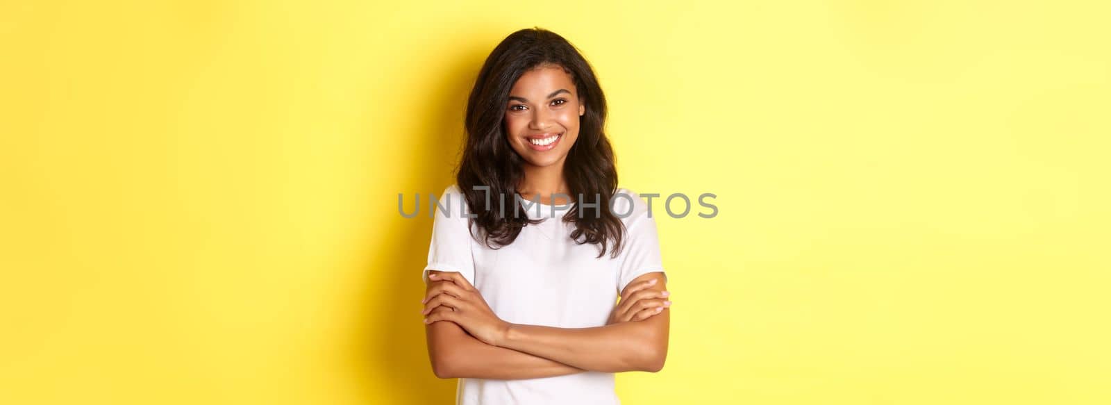 Image of happy smiling african-american girl in white t-shirt, cross arms on chest and looking confident, standing over yellow background by Benzoix