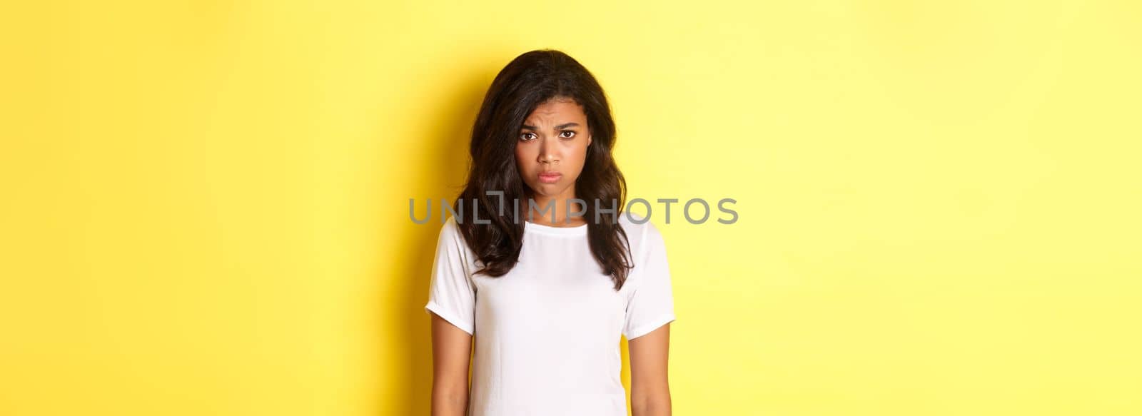 Portrait of gloomy and sad african-american girl, sulking and looking upset, standing in white t-shirt against yellow background by Benzoix