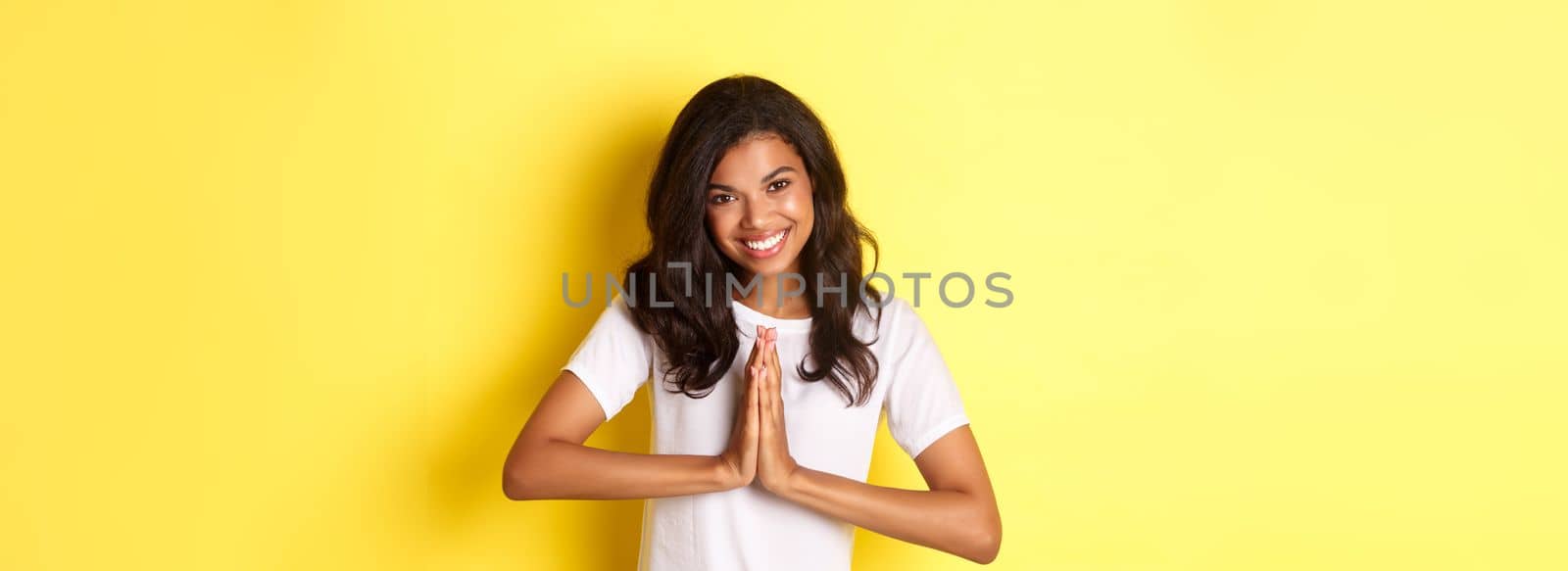 Image of cute african american woman, saying thank you and smiling, press hands together to express gratitude, standing over yellow background.