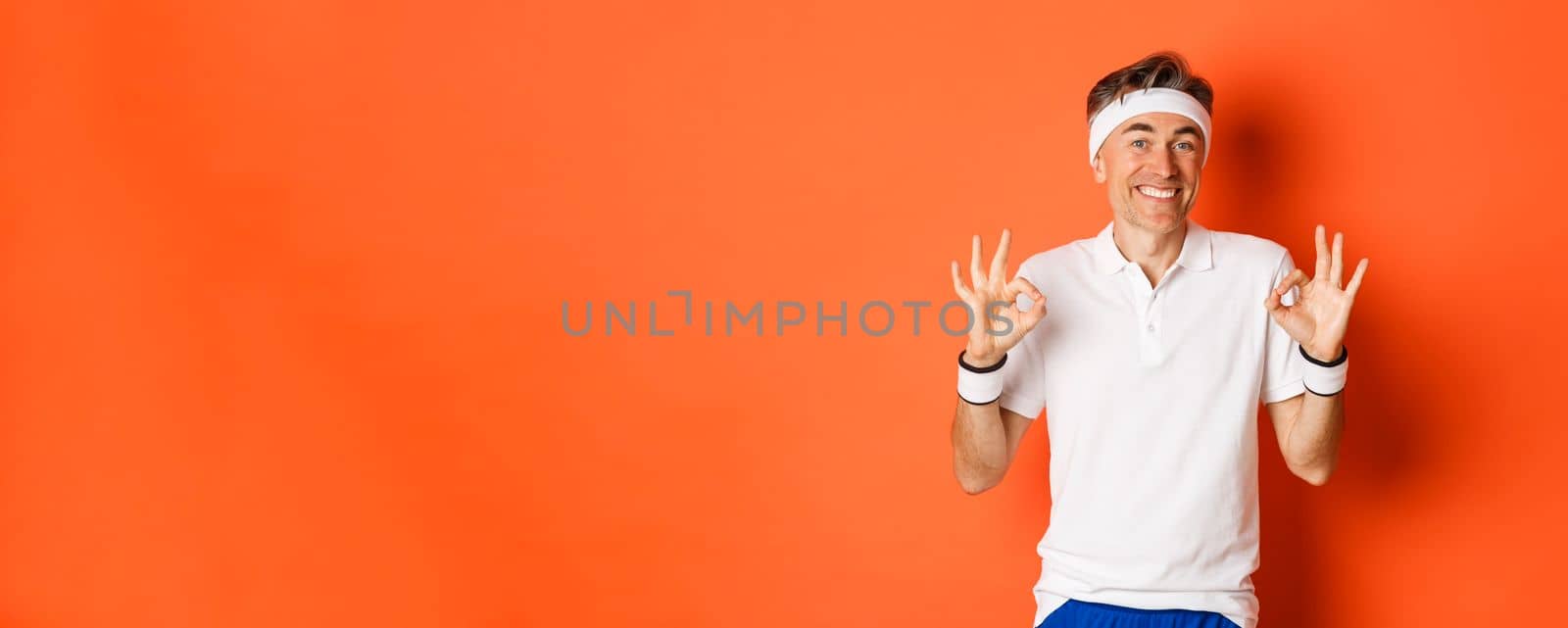 Concept of sport, fitness and lifestyle. Portrait of cheerful middle-aged male athlete, smiling pleased and showing okay signs, approve or recommend something, standing over orange background by Benzoix