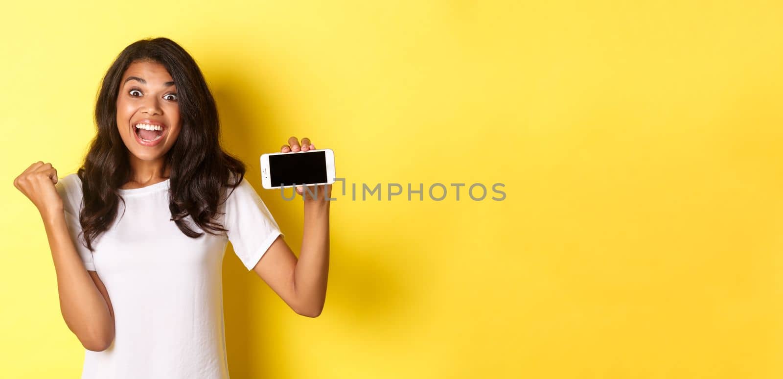 Image of beautiful african-american girl, looking excited and showing smartphone screen horizontally, standing over yellow background.