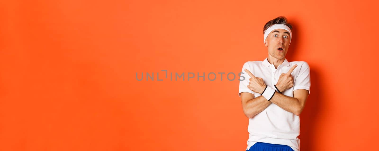 Concept of sport, fitness and lifestyle. Portrait of startled middle-aged male athlete, pointing fingers sideways and looking amazed, standing over orange background by Benzoix