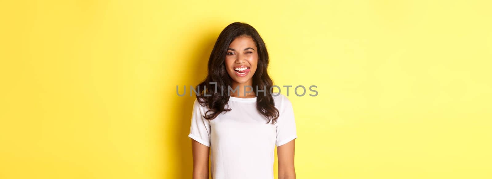 Image of carefree african-american girl in white t-shirt, showing tongue, smiling and winking happy, standing over yellow background by Benzoix