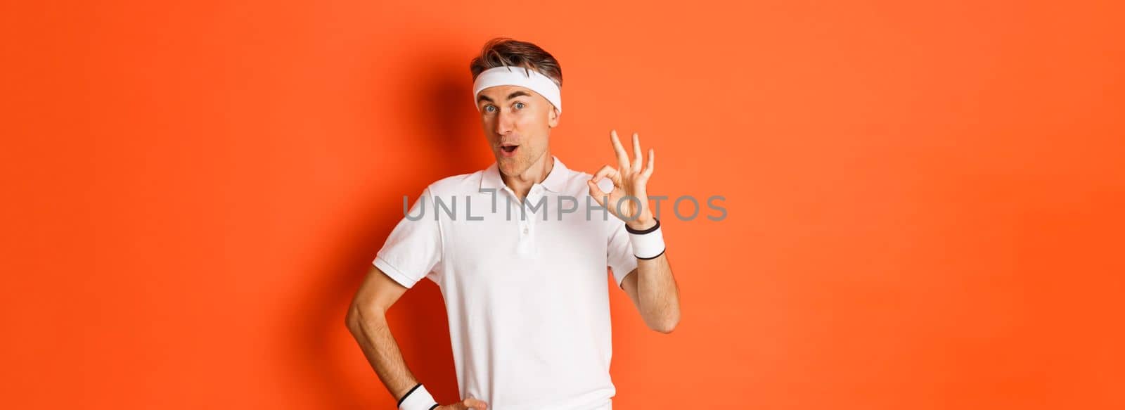 Concept of sport, fitness and lifestyle. Portrait of amazed adult man in workout uniform, showing okay sign and looking impressed, standing over orange background.