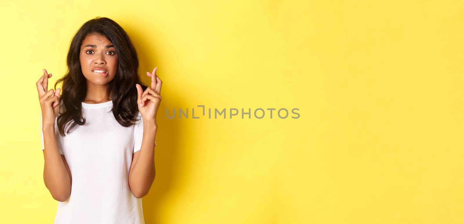 Image of anxious and hopeful african-american woman, waiting for news with fingers crossed, making wish, standing over yellow background.