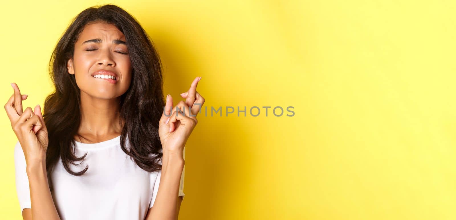 Close-up of anxious african-american woman, pleading, biting lip and close eyes while making a wish, crossing fingers for good luck, standing over yellow background.