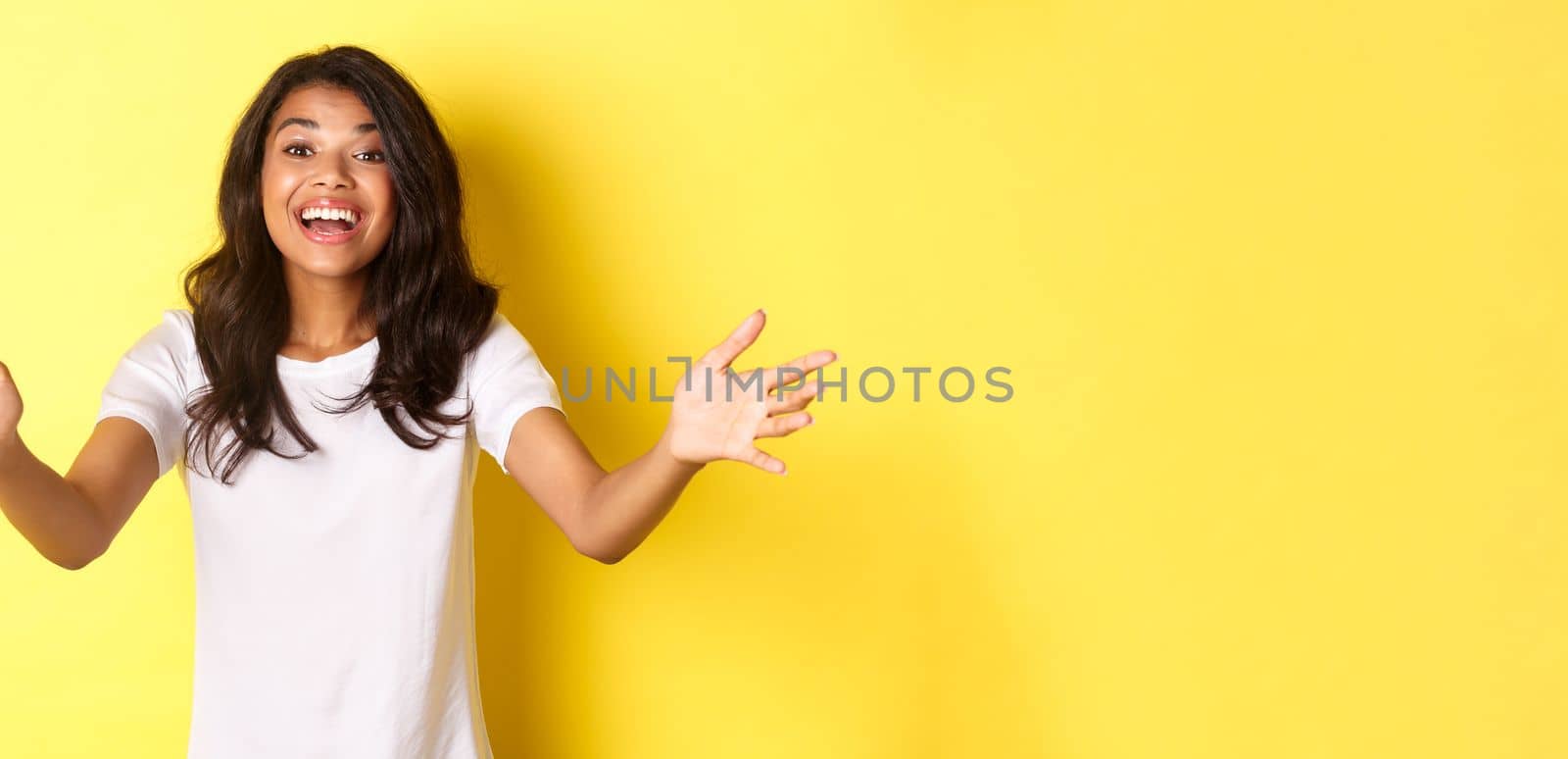 Portrait of beautiful and friendly african-american girl, reaching hands to you, wanting to hold or take something, hugging person with pleased smile, standing over yellow background by Benzoix