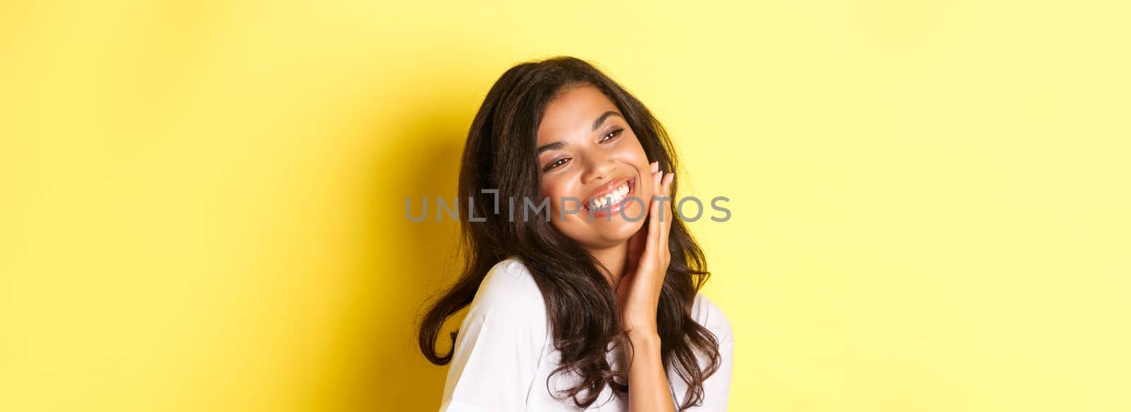 Image of gorgeous african-american woman touching her face, smiling pleased and looking left at copy space, standing over yellow background.